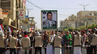 در این تصویر، گروهی از افراد در یک تظاهرات خیابانی حضور دارند. آنها لباسهای مشابهی به تن دارند و تعدادی از آنها دارای چفیه هستند. در میان جمعیت، یک پوستر بزرگ از یک شخصیت سیاسی یا نظامی به نمایش گذاشته شده است که به وضوح جلب توجه میکند. پشتزمینه تصویر ساختمانهایی با نمای شهری دیده میشود و پرچمهایی از جمله پرچم یمن نیز در دست تظاهرکنندگان مشاهده میشود. جو کلی تصویر نشاندهنده یک تجمع سیاسی است.