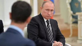 در تصویر، ولادیمیر پوتین در یک جلسه رسمی نشسته است. او لباسی تیره و رسمی بر تن دارد و به نظر میرسد در حال تفکر یا گوش دادن به سخنانی است که توسط فردی دیگر بیان میشود. زاویهای از چهرهی فرد دیگری هم در سمت چپ تصویر دیده میشود، اما مشخصات او قابل شناسایی نیست. پسزمینه تصویر به سبک رسمی و متناسب با یک جلسه سیاسی طراحی شده است.