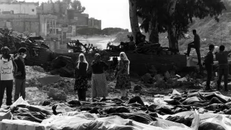 این تصویر نمایی غمانگیز و دلخراش از یک صحنهٔ بعد از بحران یا جنگ است. در جلوی تصویر، چندین جسد که با پارچهای پوشیده شدهاند، قرار دارند. در پسزمینه، گروهی از افراد حاضرند که به این صحنه نگاه میکنند، برخی از آنها اقدام به پوشاندن دهان و بینی خود کردهاند. درختان و ساختمانهای ویرانه در پشت زمینه به چشم میخورند، که حاکی از تخریب و ویرانی است. جو کلی تصویر شعاری از درد و مصیبت را منتقل میکند.