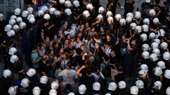 در این تصویر، گروهی از مردم در حال تظاهرات یا تجمعی هستند که با نیروهای پلیس محاصره شدهاند. افراد معترض در وسط تصویر قرار دارند و به نظر میرسد در حال گفتوگو یا گفتگو با پلیس هستند. نیروهای پلیس در اطراف آنها ایستادهاند و کلاههای مخصوصی به سر دارند. فضای تصویر نشاندهنده تنش و فعالیت اجتماعی است، با تجمع جمعیتی که به سمت نیروهای پلیس نگاه میکنند.