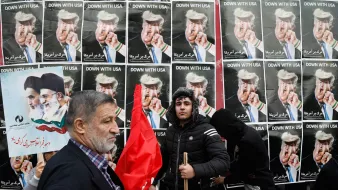 در این تصویر، گروهی از مردم در حال تظاهرات به نمایش گذاشتهاند. در پسزمینه، پوسترهایی با تصاویر رهبران سیاسی و متنی به زبان فارسی دیده میشود که به موضوعی خاص اشاره دارد. یکی از افراد به سمت دوربین نگاه میکند و فرد دیگری با یک چوب در دست و لباسی سیاه در کنار او ایستاده است. برخی از افراد دیگر نیز در این صحنه حضور دارند که نشاندهنده جو تظاهرات است. پرچمها و پلاکاردها نیز در میان جمعیت دیده میشود که نشاندهنده احساسات و پیامهای سیاسی آنها است.