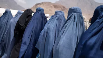 در این تصویر تعدادی زنان دیده میشوند که پوششهایی به رنگهای آبی و مشکی بر تن دارند. آنها از پشت به دوربین ایستادهاند و فقط قسمتهایی از پوششهایشان قابل مشاهده است. نمای背景 کوهها و فضای باز است، که ممکن است به منطقهای کوهستانی اشاره داشته باشد. این تصویر حس سنتی و فرهنگی خاصی را منتقل میکند.