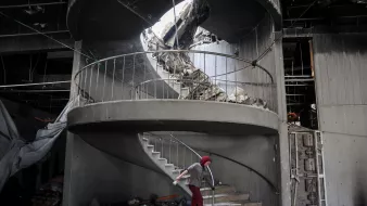 در این تصویر، یک پلهگردان دیده میشود که به دلیل تخریب، وضعیت نامناسبی دارد. بخشی از سقف بالای پلهها و دیوارها فرو ریخته و آوار زیادی در اطراف پلهها جمع شده است. یک فرد با عصا در حال حرکت در پلهها است و به نظر میرسد در تلاش است تا از این فضای خراب عبور کند. نور طبیعی از سقف خراب به داخل تابیده و فضایی دلگیر و غمانگیز به وجود آورده است.