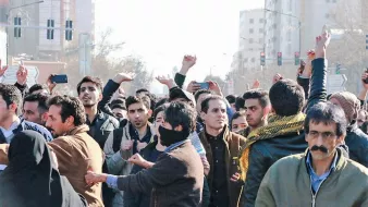 در این تصویر، گروهی از مردم در یک جمعیت انبوه دیده میشوند که به نظر میرسد در حال برگزاری یک تجمع یا اعتراض هستند. برخی از افراد دستهای خود را بالا بردهاند و دیگران به سمت دوربین نگاه میکنند. تعدادی از افراد لباسهای معمولی بر تن دارند و در پسزمینه، ساختمانها و چراغهای راهنمایی قابل مشاهده است. حالت چهرهها نشاندهندهی هیجان یا تمایل به ابراز نظر است. برخی از افراد نیز با پوشش صورت دیده میشوند.