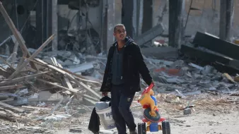 تصویر مردی را نشان میدهد که در فضایی ویران شده و پر از آوار حرکت میکند. او با ظاهری خسته، یک سطل در دست دارد و در دست دیگرش یک اسباببازی رنگی که شبیه به دوچرخه است، حمل میکند. پسزمینه تصویر شامل بقایای ساختمانها و مواد ساختمانی پراکنده است که نمایانگر وضعیت ناراحتکنندهای است.