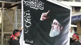 تصویر دو نفر را نشان میدهد که یک تابلو بزرگ را با تصویری از فردی در حال اشاره کردن به سمت بالا در دست دارند. پسزمینه تابلو سیاه است و بر روی آن متنهایی به زبان عربی دیده میشود. افراد حاضر در تصویر لباسهای سیاه دارند و برخی از آنها دستبندهای همرنگ بر روی سرشان دارند. این صحنه به نظر میرسد که در یک تجمع یا مراسم خاصی در حال برگزاری است.