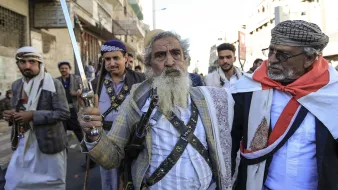 در این تصویر، چند مرد در حال راه رفتن در یک خیابان هستند. یکی از آنها، که در پیشزمینه قرار دارد، دمی بلند و ریش سفیدی دارد و یک شمشیر در دست دارد. او لباس سنتی یمنی به تن کرده و به نظر میرسد که مقام یا اهمیت خاصی دارد. دیگر مردان نیز لباسهای سنتی بر تن دارند و برخی از آنها سلاحهایی در دست دارند. جو در تصویر به نظر مهیج و پر از نشاط است و نشاندهنده یک گردهمایی یا تجمع اجتماعی میباشد.