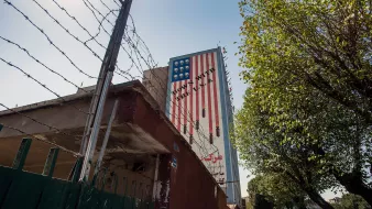 تصویر نشاندهنده دیواری است که بر روی آن پرچم آمریکا نقاشی شده است. این پرچم به گونهای طراحی شده که بخشی از آن به صورت معکوس و غیرمتعارف نمایش داده شده است. در پسزمینه، fences (حصارهایی) با میلههای تیز دیده میشود. همچنین درختان و یک پیادهرو در کنار دیوار وجود دارد. نور آفتاب به تصویر زیبایی افزوده است و فضا به طور کلی حس خاصی از تاریخ و سیاست را منتقل میکند.