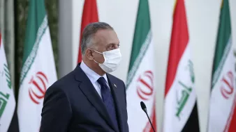 در این تصویر، مردی با روپوش رسمی و ماسک صورت دیده میشود. او در حال ایستاده و به یک سمت نگاه میکند. پشت او پرچمهای ایران و عراق در ردیف قرار گرفتهاند. فضا به نظر رسمی و جدی است، که نشاندهنده یک رویداد یا کنفرانس است.