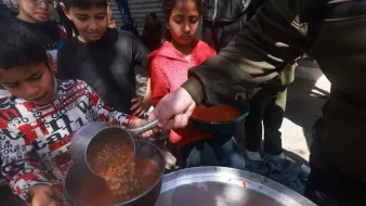 در این تصویر، گروهی از کودکان در حال دریافت غذا هستند. یکی از کودکان که لباسهای رنگارنگ دارد، با دقت در حال ریختن خورش یا سوپ به ظرفش است. در پسزمینه، سایر کودکان نیز منتظر هستند تا نوبتشان برسد. محیط بیرونی به نظر شلوغ و غیررسمی است و نشاندهنده یک فعالیت جمعی برای سرو غذا به افراد نیازمند میباشد.
