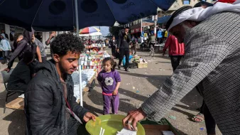 در تصویر، یک بازار محلی به چشم میخورد. در وسط تصویر، مرد جوانی نشسته و در حال فروش کالاهایی است. او یک بشقاب سبز رنگ را در دست دارد که در آن چند قلم وجود دارد. به نظر میرسد که او به مشتریاش توجه دارد که در حال انتخاب کالاهاست. در کنار آن، یک دختر بچه نیز در حال بازی کرده و به صحنه نگاه میکند. در پسزمینه، افراد دیگری در حال خرید و گشت و گذار هستند و جوی شلوغ و پرجنبوجوش به چشم میخورد. سایه چترهایی که بالای سر برخی افراد است، فضایی دلپذیر ایجاد کرده است.