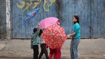 در تصویر، چند کودک در حال بازی با چترهای رنگارنگ هستند. چترها طرحهای شاد و جذابی دارند و کودکان در حالی که در دایرهای ایستادهاند، به هم نزدیک هستند. زمینه تصویر یک دیوار آبی رنگ و زمین سنگفرش شده است.