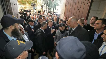 تصویر گروهی از افراد را نشان میدهد که در یک محیط شلوغ با امنیت زیاد در حال گفتگو هستند. تعدادی از افراد یونیفورمپوش به نظر میرسد که نگهبان هستند و در حال محافظت از یک فرد دیگر هستند. افراد دیگر در اطراف آنها ایستادهاند و به نظر میرسد که برخی از آنها مشغول ضبط یا عکاسی از این صحنه هستند. فضا به شدت پر ازدحام و حاکی از توجه رسانهای است.