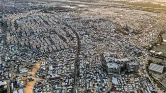 این تصویر نمای هوایی از یک منطقه شلوغ و پرجمعیت است. در اینجا تعداد زیادی ساختمان و خانه های کوچک دیده میشود که در کنار هم قرار گرفتهاند. خیابانهایی که به نظر میرسد مردم در آن تردد دارند، میانه تصویر را تقسیم کردهاند. همچنین، در برخی نواحی، چادرها و سازههای موقت دیده میشوند که نشاندهنده زندگی در شرایط سخت و غیرفرهنگین است. این تصویر، وضعیتی را به نمایش میگذارد که بیانگر چالشهای اجتماعی و اقتصادی است.