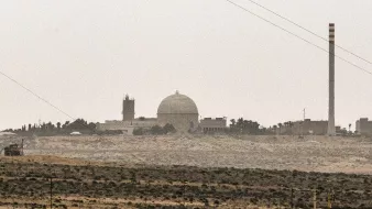 تصویر نمایی از یک تاسیسات صنعتی یا علمی را نشان میدهد که دارای یک گنبد بزرگ و چند ساختمان دیگر است. در سمت راست تصویر، یک دودکش بلند به چشم میخورد که ممکن است مربوط به فعالیتهای صنعتی باشد. فضای اطراف تصویر سرسبز نیست و بیشتر به بیابان یا زمینهای خشک مشابهت دارد. آسمان نیز ابری و کدر به نظر میرسد. این تصویر ممکن است به یک مرکز تحقیقاتی یا نیروگاه اشاره داشته باشد.