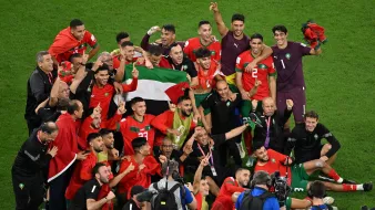 این تصویر نشاندهنده خوشحالی و شادی تیم ملی فوتبال مراکش پس از یک پیروزی بزرگ است. بازیکنان و اعضای کادر فنی تیم در حال جشن گرفتن هستند و بعضی از آنها پرچم مراکش را در دستان خود دارند. فضای تصویر پر از انرژی مثبت و شادی است و بازیکنان با لبخند و هیجان در کنار یکدیگر قرار دارند. همچنین تعدادی از عکاسان و اعضای رسانه نیز در حال تصویربرداری از این لحظه مهم هستند.