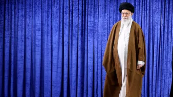 در تصویر، فردی با لباس سنتی روحانی به رنگهای قهوهای و سفید دیده میشود. او کلاهی مشکی بر سر دارد و ریش بلندی دارد. پسزمینه تصویر شامل پردهای به رنگ آبی است. فرد در حال حرکت به سمت سمت راست تصویر قرار دارد. این تصویر به طور کلی حس وقار و احترام را القا میکند.