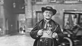 این تصویر یک مرد با لباسهای سنتی و رسمی را نمایش میدهد. او کلاهی بزرگ بر سر دارد و لباسش بلند و مشکی است که با دکمههای درخشانی تزئین شده است. مرد در حال ایستادن بر روی سنگفرش خیابانی است و در پسزمینه، ساختمانهای قدیمی و یک خودروی تاریخی به چشم میخورد. به نظر میرسد او شخصیتی مهم یا مذهبی باشد. نور و کنتراست تصویر موجب برجسته شدن جزئیات لباس و چهرهاش شده است.