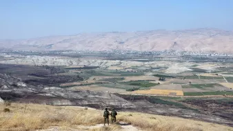در این تصویر، دو نفر در حال ایستادن بر روی یک تپه یا ارتفاع هستند و به منظرۀ وسیع و زیبای زیر پای خود نگاه میکنند. در زیر، دشتها و زمینهای کشاورزی با رنگهای مختلف دیده میشوند. دامنههای کوهها در دوردست قرار دارند و آسمان آبی و صاف به چشم میخورد. فضا به نظر آرام و دلنشین میرسد و نشاندهندهٔ مناظر طبیعی و انسانی در منطقهای کوهستانی است.