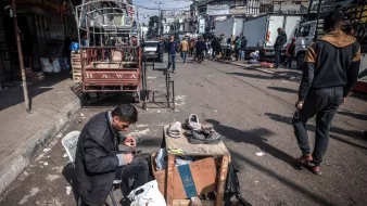 در تصویر، مردی در حال تعمیر یا واکس زدن کفشها است. او روی یک صندلی نشسته و وسایل کارش را بر روی یک میز کوچک چوبی قرار داده است. در اطراف او، افرادی در حال عبور و مرور هستند و تعدادی خودروی باربری در پسزمینه دیده میشود. فضایی شلوغ و شهری به نظر میرسد و فعالیتهای روزمره در حال انجام است. چند کشیده و بقیه اجناس نیز در اطراف قرار دارند.