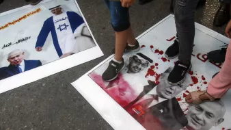 در این تصویر، تعدادی از افراد با پا بر روی پوسترهایی که عکسهایی از شخصیتهای سیاسی معروف را نشان میدهند، ایستادهاند. یکی از پوسترها به تصویر نتانیاهو مربوط میشود و دیگری شامل تصاویری از ترامپ و یک شخصیت دیگر است. نشانهای خون و نوشتهها بر روی پوسترها به وضوح دیده میشوند و این تصویر به نظر میرسد در یک تظاهرات یا اعتراض گرفته شده باشد.