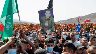 در تصویر، گروهی از افراد در یک تظاهرات حضور دارند. تعداد زیادی از افراد پرچمهایی به رنگهای مختلف در دست دارند. یکی از تظاهرکنندگان تصویری از یک مرد را بالا نگه داشته است. بیشتر افراد ماسک به صورت دارند و در پسزمینه، زمینهای سرسبز و تپههایی دیده میشود. به نظر میرسد که این تجمع به منظور اعتراض یا یادبود برگزار شده باشد.