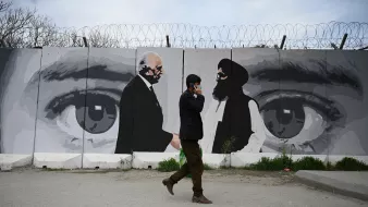 در این تصویر یک مرد در حال راه رفتن در کنار دیواری با یک نقاشی بزرگ است. دیوار دارای تصاویری از دو مرد است که به نظر میرسد در حال گفتوگو هستند. این نقاشی شامل چشمان بزرگی است که به وضوح بر روی دیوار کشیده شده و احساس عمیقی از توجه و نظارت را منتقل میکند. محیط اطراف خیابانی است و مرد به نظر میرسد مشغول صحبت کردن با تلفن همراهش باشد. طراحی و رنگها نسبتاً ملایم و معاصر به نظر میرسند.