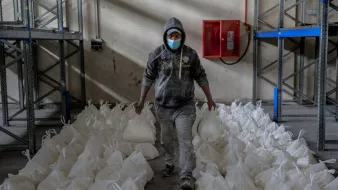 در این تصویر، یک مرد با پوشش خاکستری و کلاهی بر سر، در حال حرکت در یک محل انبار است. او یک ماسک به صورت دارد و در دو دست خود کیسههای سفیدی را حمل میکند. اطراف او ردیفهایی از کیسههای مشابه به طور منظم روی زمین قرار دارند. دیوارها و قفسههای فلزی در پسزمینه قابل مشاهده هستند که به نظر میرسد برای نگهداری کالاها استفاده میشوند. نور کمی در فضا وجود دارد و جو عمومی، فعالیتی را در زمینه توزیع یا انبارداری نشان میدهد.