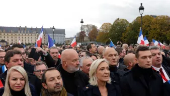 در این تصویر، گروهی از مردم در یک تجمع یا راهپیمایی در فضای باز به نمایش درآمدهاند. بسیاری از افراد پرچمهای فرانسه را در دست دارند. در میان جمعیت، افرادی با چهرههای مختلف دیده میشوند که نشاندهنده نظرات و احساسات مختلفی هستند. برخی از افراد لباسهای رسمی و گرم پوشیدهاند و به نظر میرسد موضوع این تجمع سیاسی باشد. آسمان ابری است و فضای عمومی کمی شلوغ و پرتحرک به نظر میرسد.