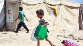 در تصویر، پسربچهای دیده میشود که در حال حمل یک کولهپشتی به رنگ صورتی و یک ظرف سبز رنگ است. او در یک محیط بیابانی و خشک قرار دارد که چادرهای موقتی در پسزمینه به چشم میخورد. پسربچه لباسی ساده و راحت پوشیده و به نظر میرسد که در حال حرکت به سمت مقصدی است. در کنار او، کودک دیگری نیز در حال گذر است. فضای تصویر حالتی از زندگی در شرایط سخت را به نمایش میگذارد.