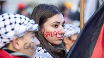 در تصویر، چند نفر در حال شرکت در یک تجمع یا اعتراض به چشم میخورند. یک دختر جوان با چهرهای جدی و نگران، روی صورتش نوشتهای به رنگ قرمز دارد که "Gaza" به نمایش گذاشته شده است. او همچنین شال یا لباسی با طرحهای خاص به تن دارد. در کنار او، پسرکی نیز دیده میشود که روی صورتش نوشتهای مشابه به رنگ سیاه دارد. فضای کلی تصویر نشاندهنده شور و نشاط در حمایت از مسئلهای است که به نظر میرسد به غزه مربوط میشود. دیگر افراد نیز در پسزمینه حضور دارند و فضا به وضوح یک تجمع اجتماعی را تداعی میکند.