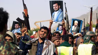 در این تصویر، گروهی از مردم در حال برگزاری یک تجمع هستند. تعدادی از شرکتکنندگان سلاحهای خود را بلند کردهاند و تعدادی دیگر تصاویری از یک فرد را در دست دارند. این تجمع به نظر میرسد که از جنبه سیاسی و اجتماعی برخوردار باشد. مردمی که در تصویر دیده میشوند، لباسهای سنتی و محلی به تن دارند و برخی از آنها روسریهایی به سر بستهاند. پسزمینه تصویر شامل تعدادی سازه و فضای باز است.