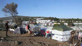 تصویر نشاندهنده یک منطقه با وضعیت نامناسب است که در آن چادرهایی به عنوان محل سکونت نصب شدهاند. در پسزمینه، زمینهای ناهموار و درختانی مشاهده میشود. پسزمینه شهری با ساختمانهایی دیده میشود که نسبتاً به چادرها نزدیک هستند. احساس کلی تصویر بیانگر شرایط سخت زندگی افرادی است که در این چادرها سکونت دارند. آسمان آبی و روز روشن است، اما وضعیت محیط نشاندهنده فقر و مشکلات معیشتی است.