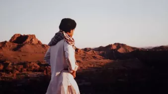 در این تصویر، فردی با لباس سنتی و شال بر دوش، به سمت یک چشمانداز طبیعی نگاه میکند. زمینه تصویر شامل کوههای بلند و دشتهای وسیع است که نور ملایم غروب خورشید بر آنها تابیده شده است. رنگهای گرم غروب و سایههای بلند کوهها زیبایی خاصی به صحنه دادهاند. احساس آرامش و سکوت در فضای تصویر به وضوح حس میشود.