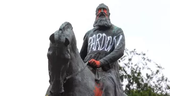 این تصویر نشاندهنده یک مجسمه است که یک شخصیت سوار بر اسب را به تصویر کشیده است. مجسمه به نظر میرسد که به طور هنری با رنگهای مختلف نقاشی شده و روی آن عبارتهایی نوشته شده است. کلمات "PARDON" و "RACIST" به وضوح روی مجسمه دیده میشوند، که بیانگر اعتراضات یا نظرات انتقادی نسبت به این شخصیت تاریخی است. زمینه تصویر شامل درختان و آسمان ابری است.