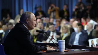 در این تصویر، ولادیمیر پوتین، رئیسجمهور روسیه، در حال سخنرانی یا پاسخ به سؤالات در یک کنفرانس مطبوعاتی به نظر میرسد. او در حال نشستن در کنار یک میز است که میکروفون دارد و در مقابل او تعدادی از خبرنگاران و حضار به نظر میرسند. محیط اطراف کمی تاریکتر است و تمرکز اصلی بر روی پوتین قرار دارد. تعدادی از افراد حاضر در جمع در پسزمینه دیده میشوند.