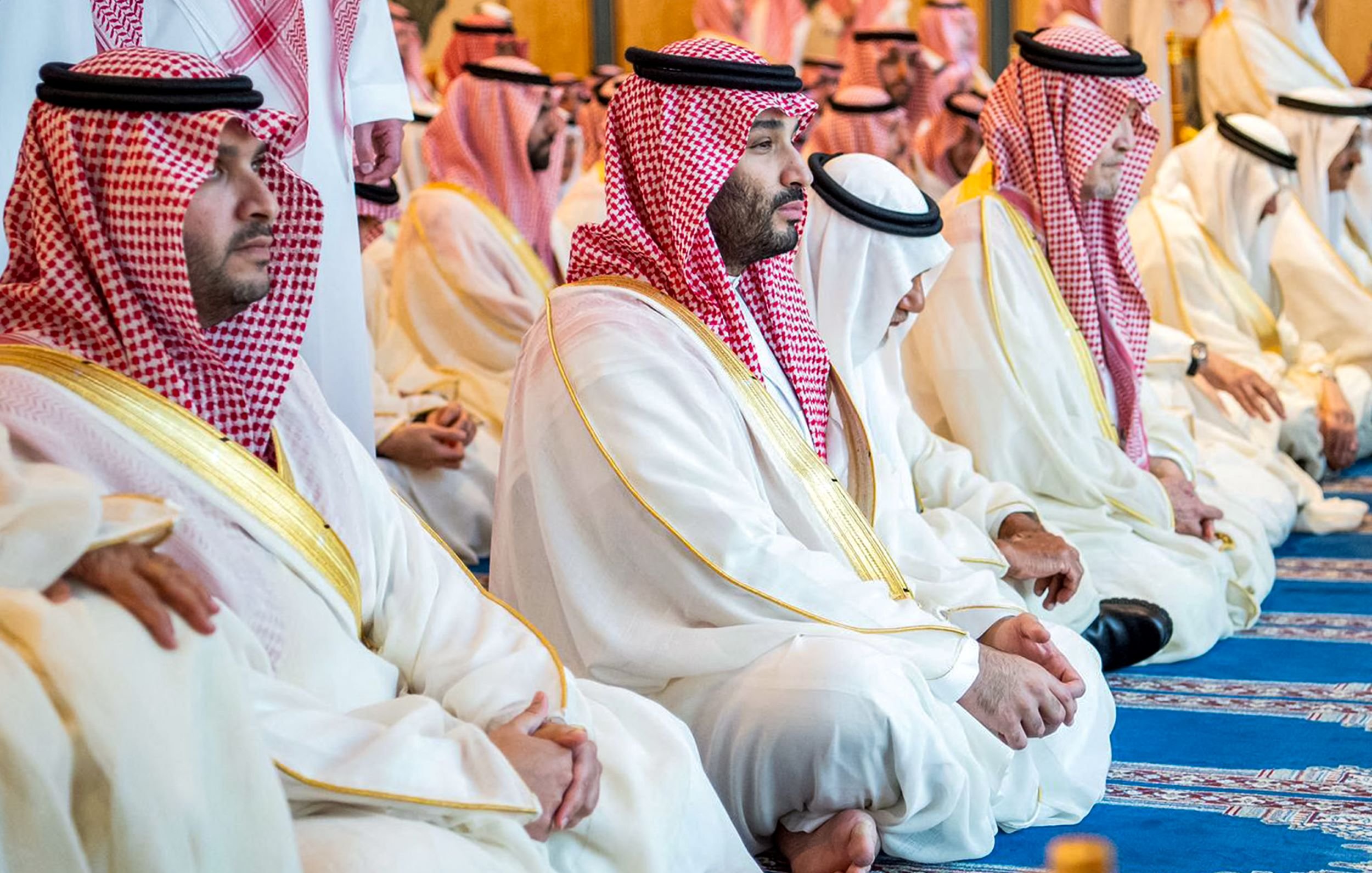 L'image montre un groupe d'hommes assis, probablement en train de prier ou de méditer. Ils portent des vêtements traditionnels saoudiens, comme des thobes blancs et des chapeaux appelés ghutras, souvent ornés de bandes rouges et blanches. L'atmosphère semble empreinte de sérénité et de respect, reflet d'un moment spirituel partagé. Le décor autour d'eux est élégant et peut inclure des éléments architecturaux typiques d'un lieu de culte.