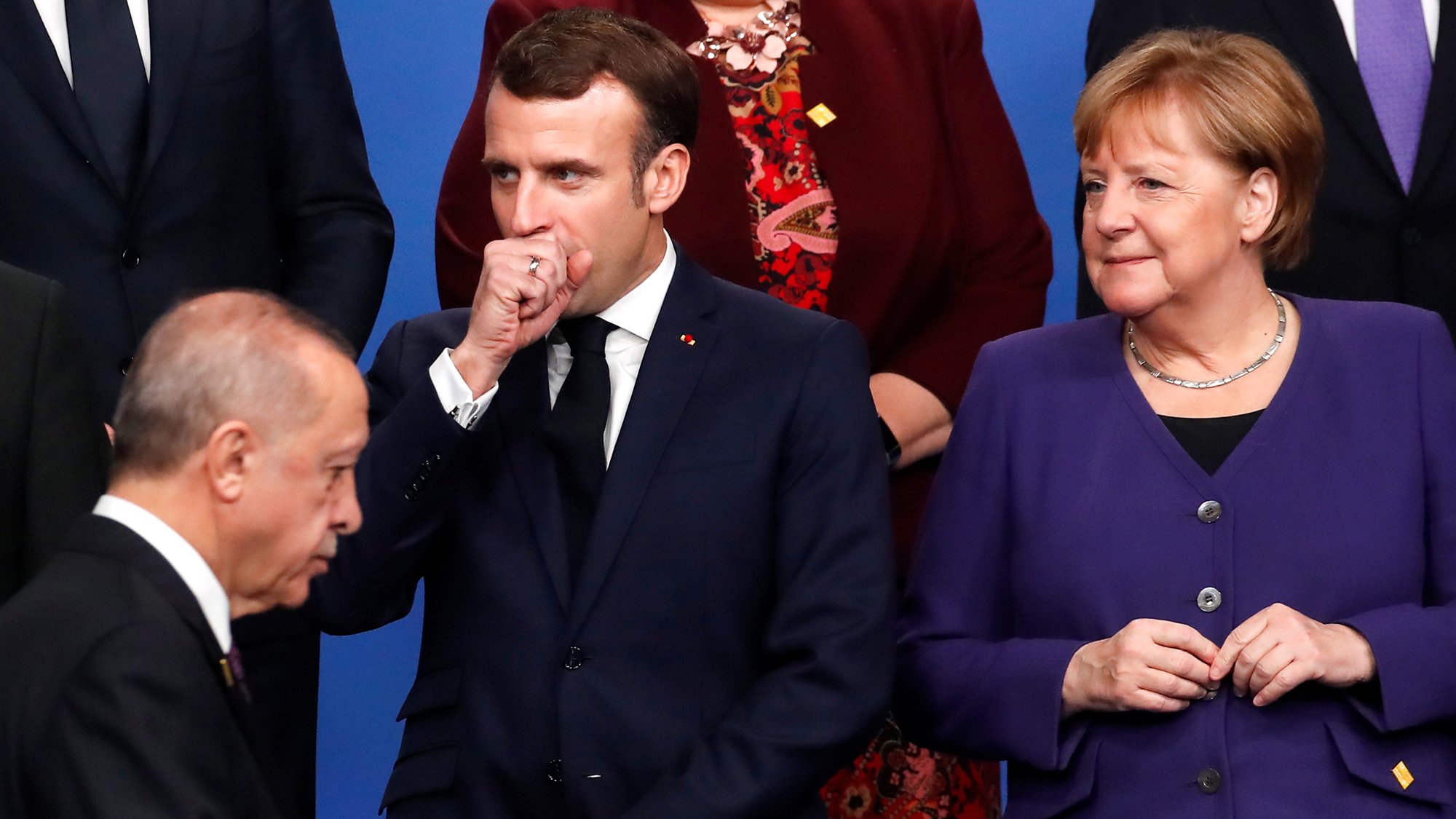 L'image montre un groupe de dirigeants politiques rassemblés lors d'un événement officiel. Au premier plan, on peut voir un homme qui se tient à gauche, probablement en train de parler ou d'écouter. Au centre, un homme en costume sombre semble réfléchir, avec une main devant sa bouche. À droite, une femme porte un vêtement violet et observe la scène avec un léger sourire. L'atmosphère semble être formelle, typique des rencontres diplomatiques.