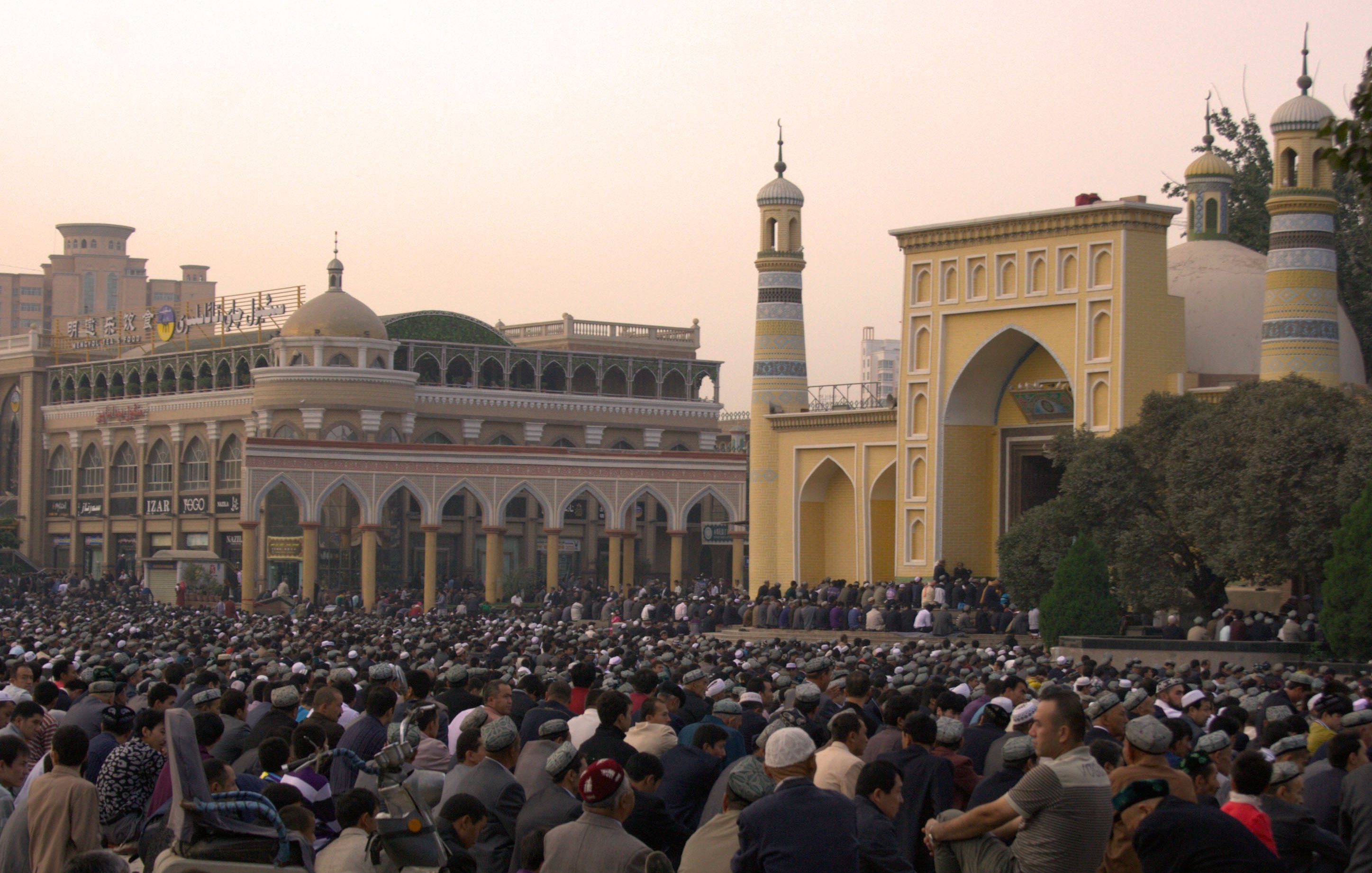 L'image montre une grande foule de personnes rassemblées dans une zone en plein air, probablement pour un service religieux. À l'arrière-plan, on aperçoit des bâtiments architecturaux impressionnants, avec des minarets et des ornements qui évoquent une ambiance culturelle riche. Les gens sont principalement assis, attentifs à ce qui se passe, créant une atmosphère de commémoration ou de prière collective. La lumière du soir donne une teinte chaleureuse à la scène.