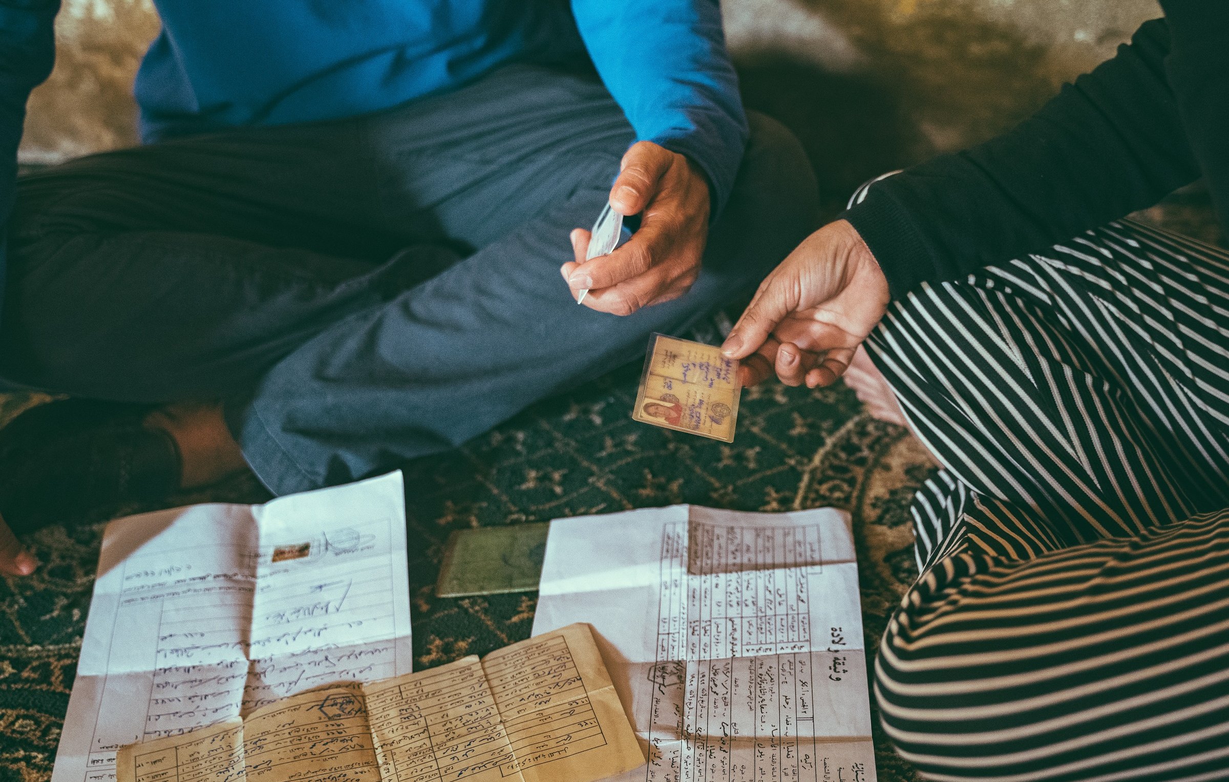L'image montre deux personnes assises par terre, entourées de divers documents étalés devant elles. L'une des personnes tient une carte d'identité ou un document similaire, tandis que l'autre semble utiliser un marqueur pour écrire ou annoter quelque chose. Les documents sur le sol sont variés, comprenant des papiers manuscrits et des feuilles à imprimer. L'environnement semble intime et informel, suggérant une discussion importante ou une transaction en cours.