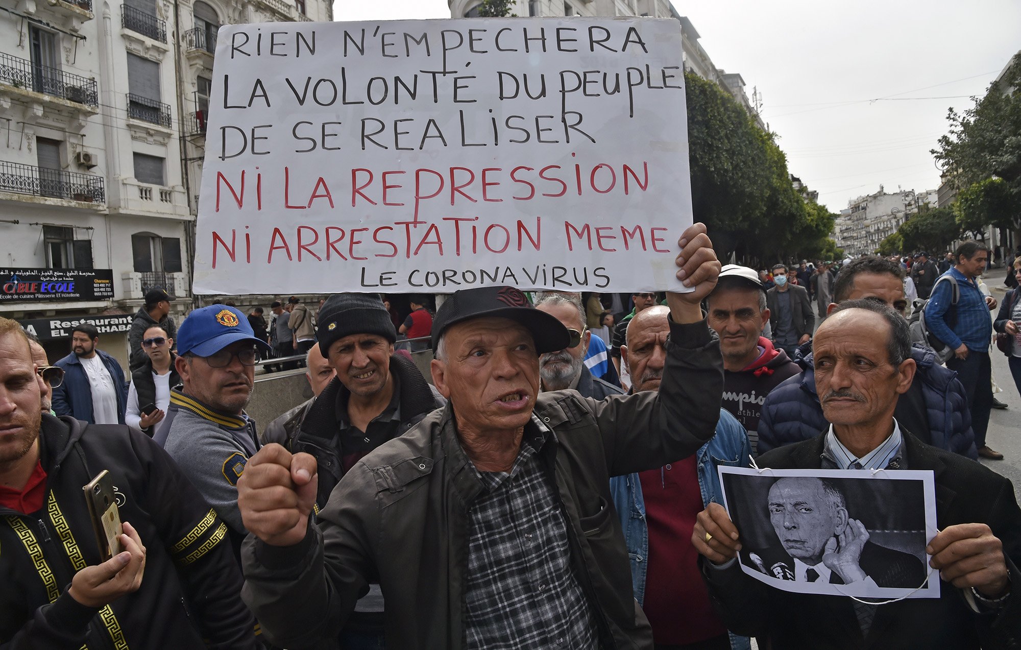 Sur l'image, un groupe de personnes se rassemble dans une rue, probablement lors d'une manifestation. Un homme tient une grande pancarte sur laquelle est écrit : "Rien n'empêchera la volonté du peuple de se réaliser ni la répression ni arrestation même le coronavirus". L'assemblée semble déterminée, et des expressions sérieuses sont visibles sur les visages des manifestants. L'ambiance évoque une lutte pour des droits ou des changements sociaux, sur fond de contestation.