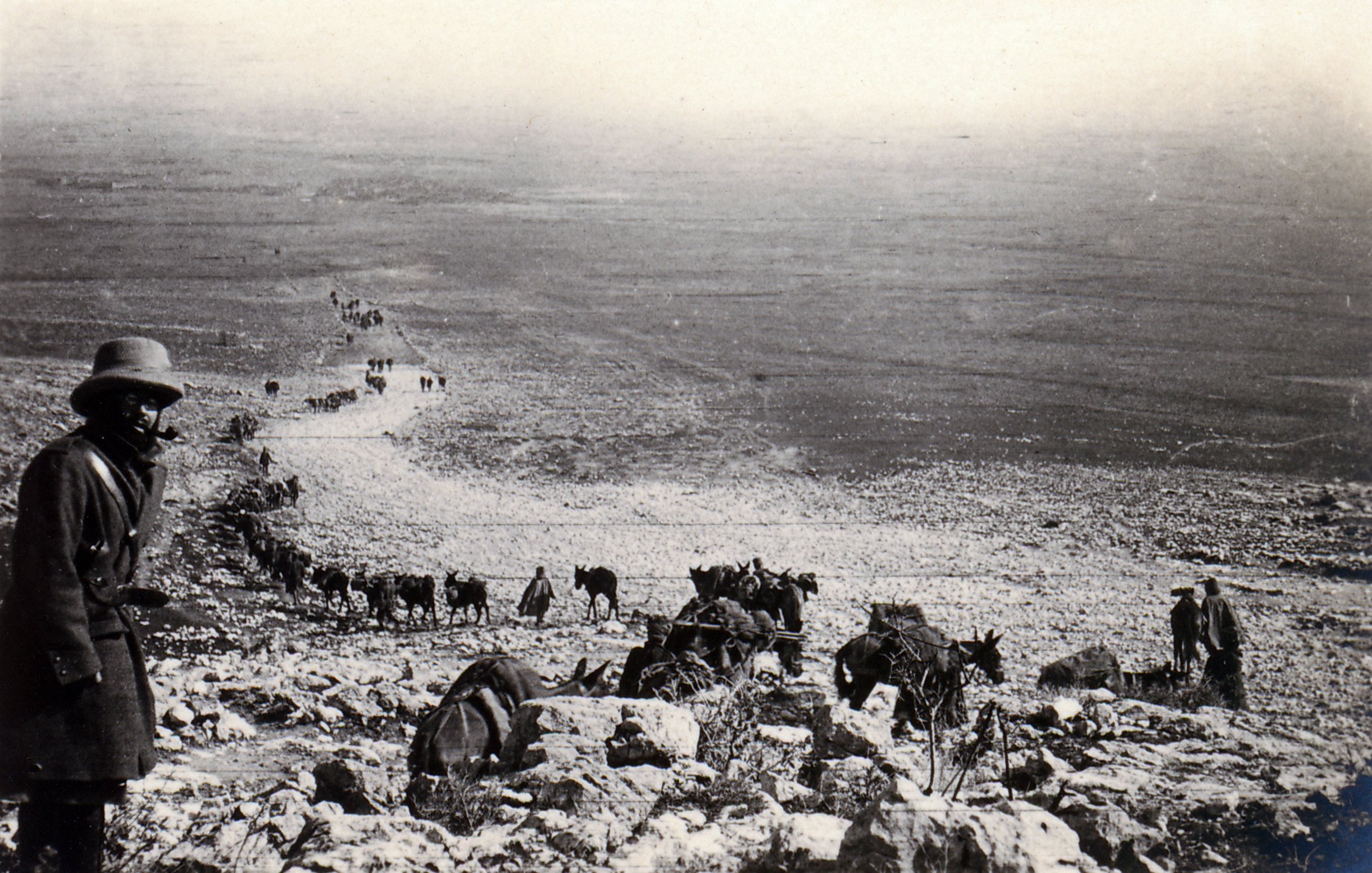 L'image montre un paysage désertique avec un groupe de personnes et d'animaux, probablement des mulets ou des ânes, qui avancent sur un chemin rocailleux. À l'avant, un homme en manteau et en chapeau observe la scène. Au loin, on peut voir une longue file de silhouette se déplaçant à travers des terrains arides. L'atmosphère est presque nostalgique, évoquant un temps passé.