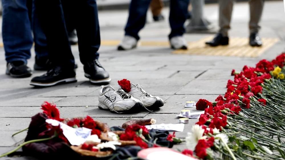 L'image montre un sol avec une paire de chaussures de sport blanches laissées à l'écart. Autour des chaussures, il y a une traînée de fleurs rouges, probablement des œillets, qui sont disposées en ligne. On peut également voir d'autres objets sur le sol, tels que des photos et des éléments décoratifs. En arrière-plan, des gens sont visibles, mais leur apparence et leurs détails ne sont pas clairement discernables. L'ensemble de la scène dégage une atmosphère de mémoire ou de commémoration.