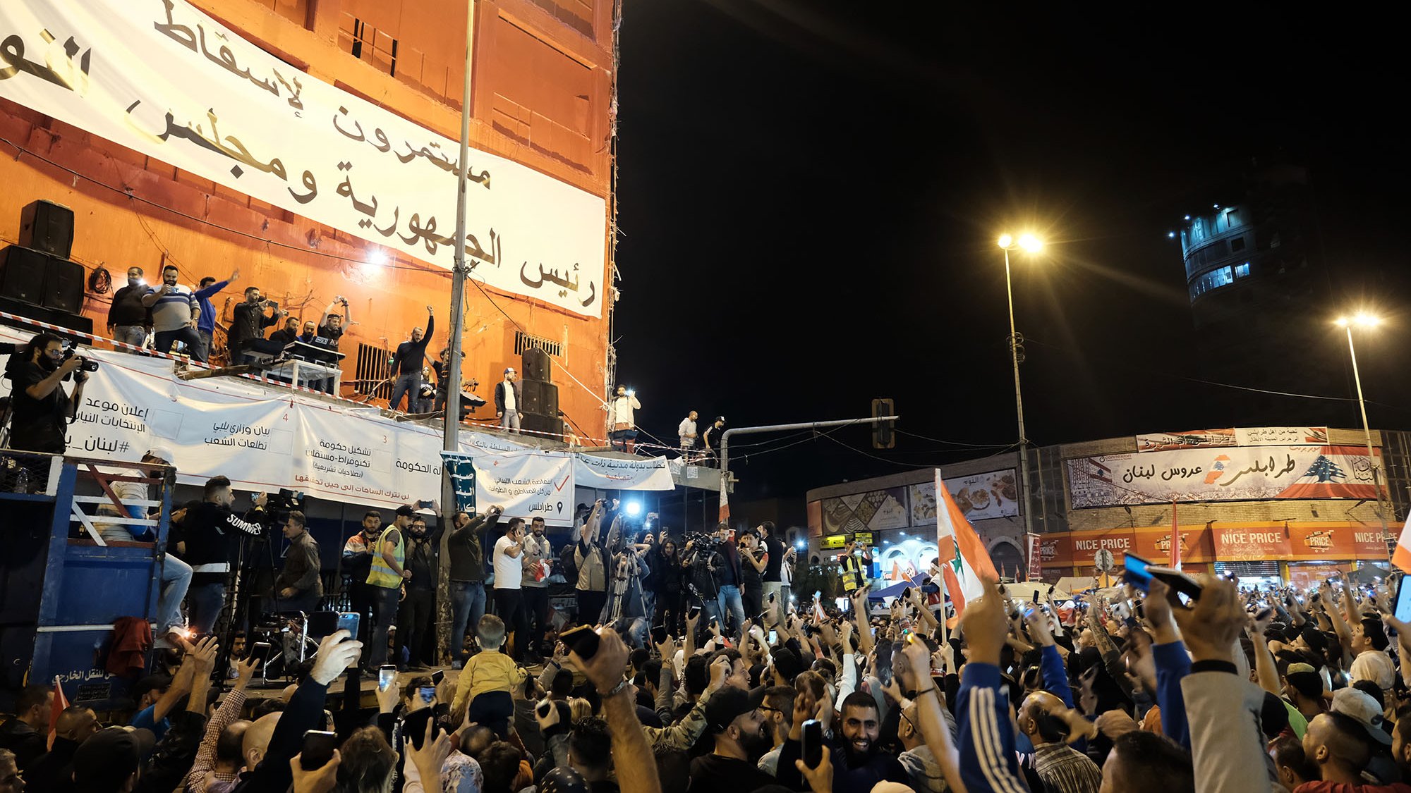 L'image montre une grande foule rassemblée dans une place publique, manifestant avec enthousiasme. Les gens lèvent les mains et brandissent des drapeaux, exprimant des émotions fortes. À l'arrière-plan, on peut voir un grand panneau affichant des messages politiques. Des personnages sont visibles sur une scène surélevée, semblant s'adresser à la foule. L'ambiance semble festive et dynamique, avec des lumières qui illuminent la scène.