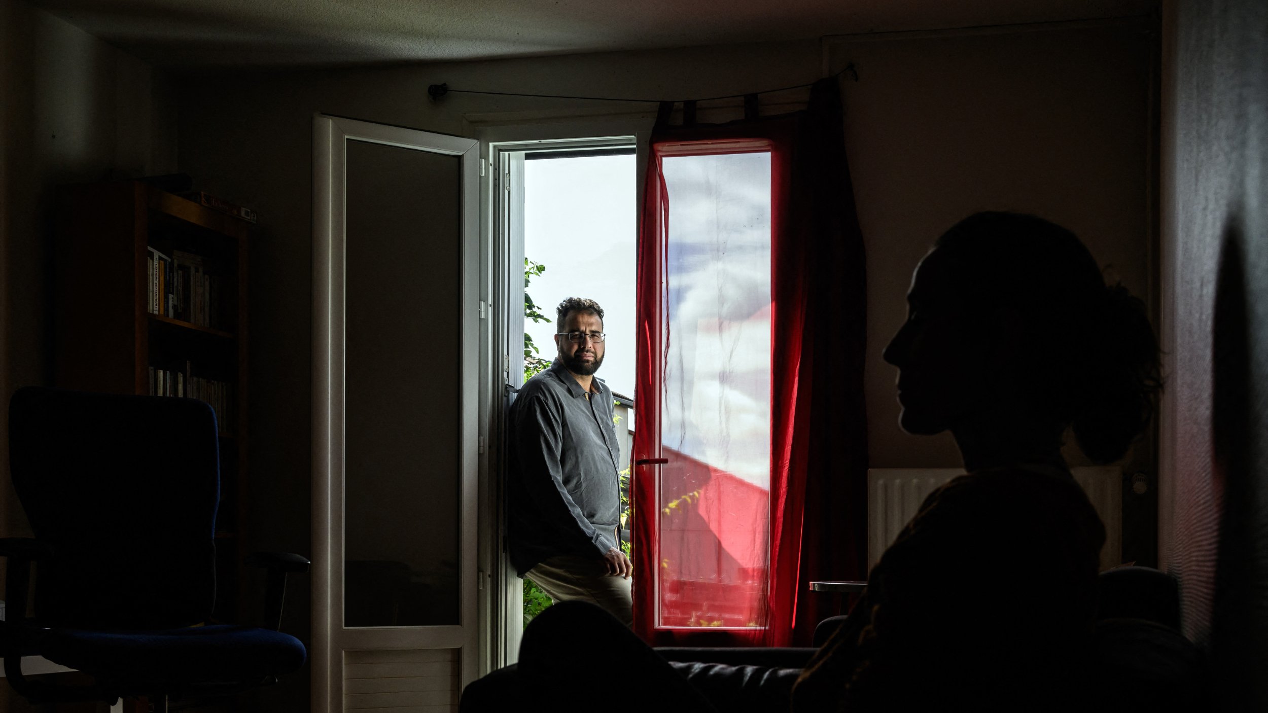 L'image montre un intérieur de maison ou d'appartement. Au premier plan, on voit la silhouette d'une personne assise sur un canapé, éclairée par une lumière douce, tandis qu'en arrière-plan, un homme est debout près d'une porte ouverte qui donne sur l'extérieur. La lumière entre par la fenêtre de façon à créer un contraste intéressant entre les deux personnages. Les éléments de la pièce, tels qu'un fauteuil et des étagères, ajoutent à l'ambiance chaleureuse et intime de la scène.