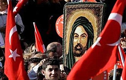 L'image montre une foule rassemblée lors d'une manifestation. Au centre, une personne tient une grande image représentant un personnage avec une longue barbe et un turban, entourée de drapeaux turcs. La foule semble engagée et énergique, avec des expressions variées sur les visages des participants.