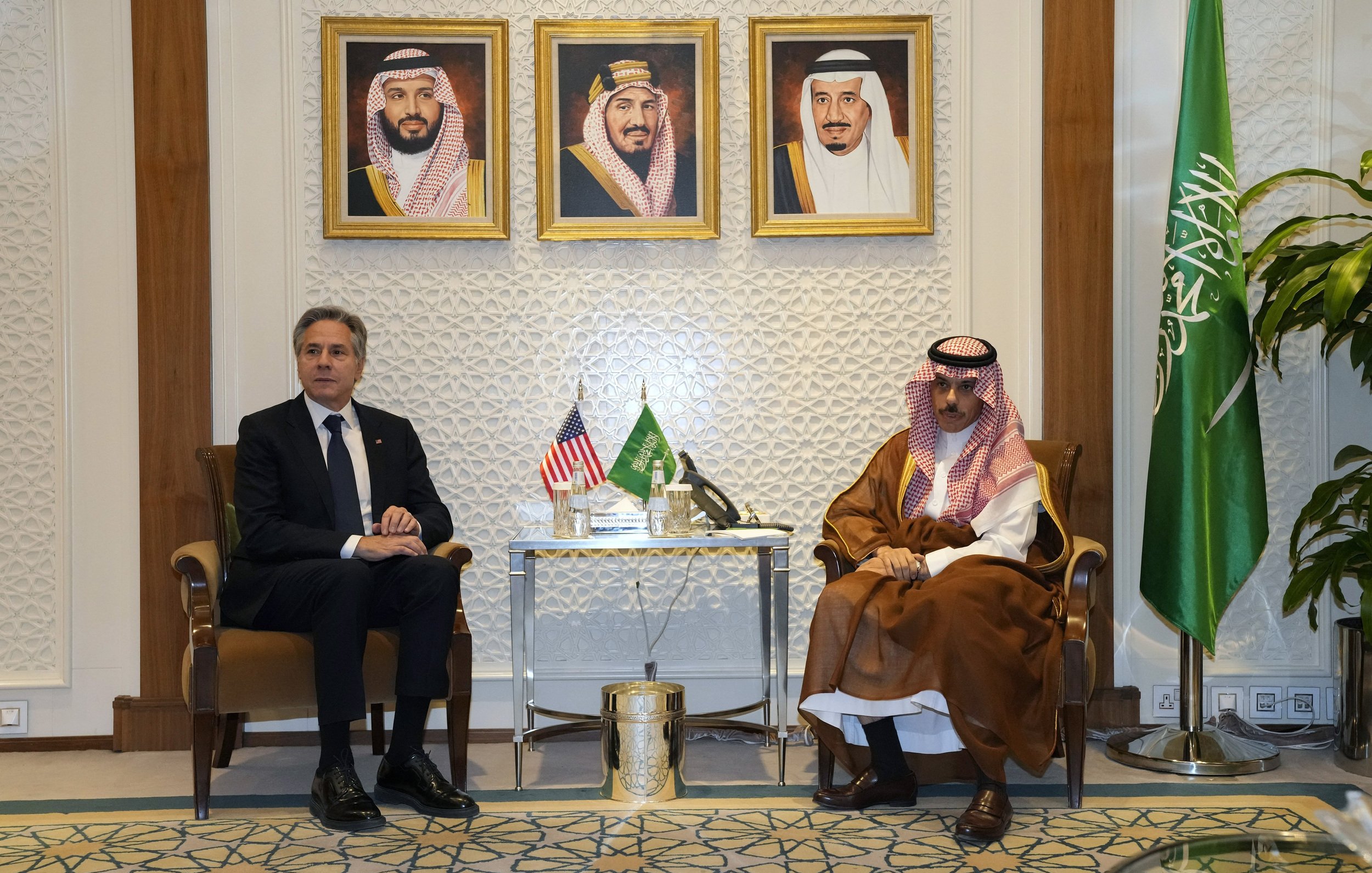 L'image montre deux hommes assis côte à côte dans un cadre formel. À l'arrière-plan, il y a trois portraits encadrés de dirigeants saoudiens. Sur une table entre eux, se trouvent des éléments décoratifs. L'un des hommes porte un costume sombre et l'autre est habillé en tenue traditionnelle saoudienne. À droite, on aperçoit un drapeau saoudien. L'atmosphère semble officielle et diplomatique.