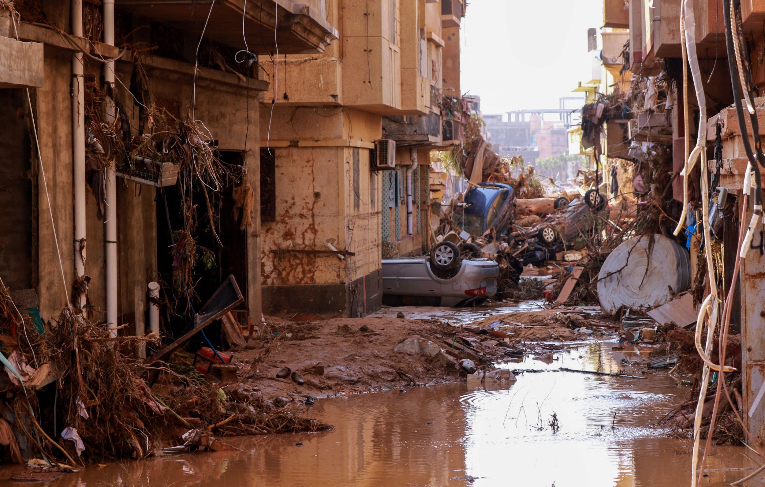L'image montre une rue dévastée par une inondation. On peut voir des bâtiments endommagés, avec des débris et de la boue accumulés sur le sol. Des voitures sont renversées et se trouvent parmi les débris, tandis qu'une faible couche d'eau stagnante reflète la destruction environnante. L'atmosphère est sombre, soulignant les conséquences dramatiques de la catastrophe.