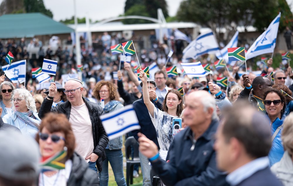 L'image montre une grande foule rassemblée, tenant des drapeaux israéliens et sud-africains. Les personnes semblent engagées et heureux, certaines souriant et levant les drapeaux en signe de soutien. L'environnement est un espace extérieur, et on peut voir des stands ou des tribunes en arrière-plan, suggérant un événement public ou une cérémonie. L'atmosphère semble festive et solidaire.