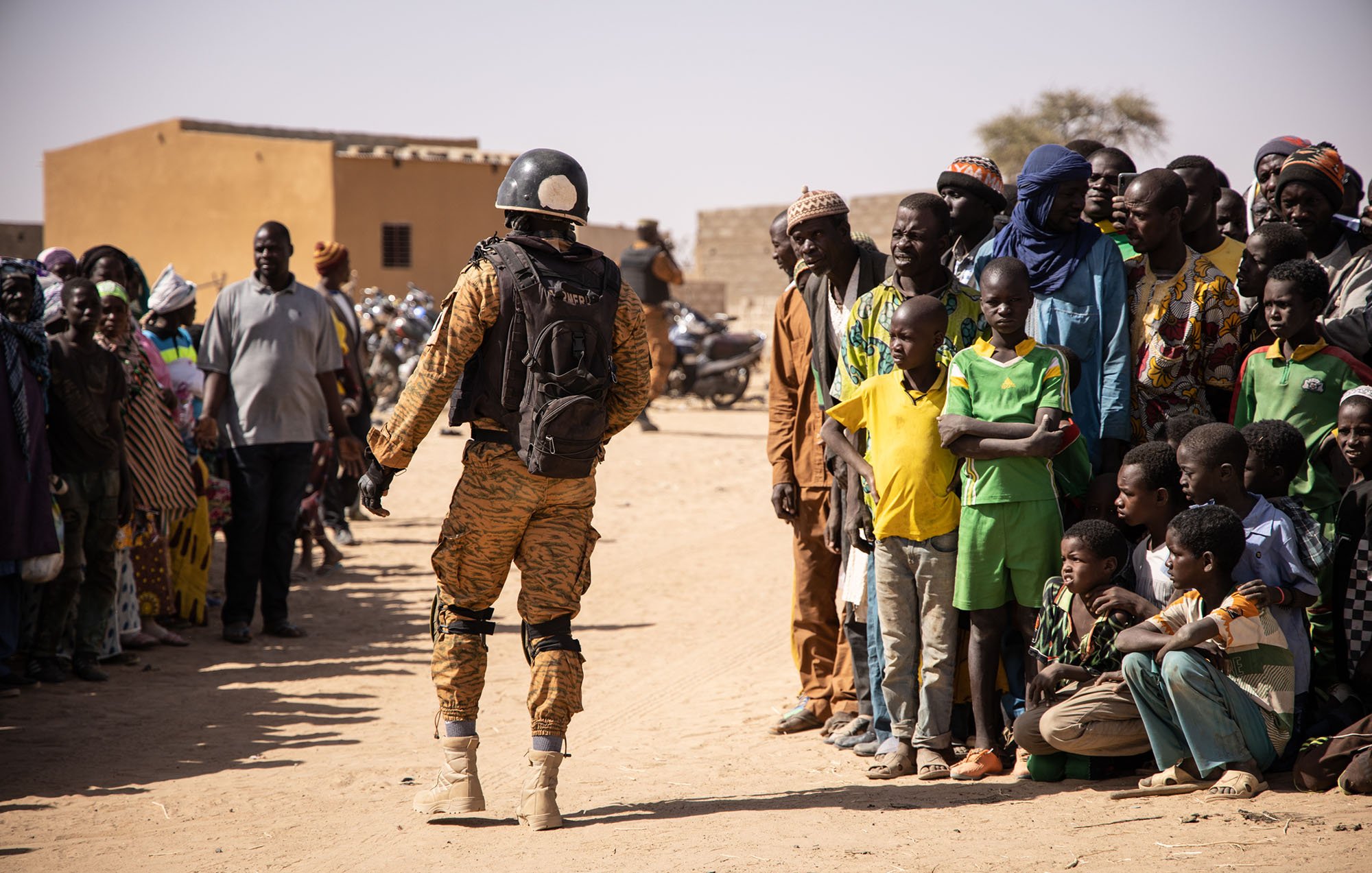 L'image montre un soldat en uniforme militaire, portant un casque et un sac à dos, se tenant face à une foule. Les personnes dans la foule sont de différentes tranches d'âge, allant des enfants aux adultes, et semblent observer le soldat avec attention. Le décor évoque une ambiance de village rural, avec des bâtiments simples et un sol en terre battue. Les couleurs de l'uniforme du soldat contrastent avec celles des vêtements des membres de la foule. L'atmosphère générale semble être celle d'une interaction, peut-être dans le cadre d'une mission de sécurité ou d'assistance.